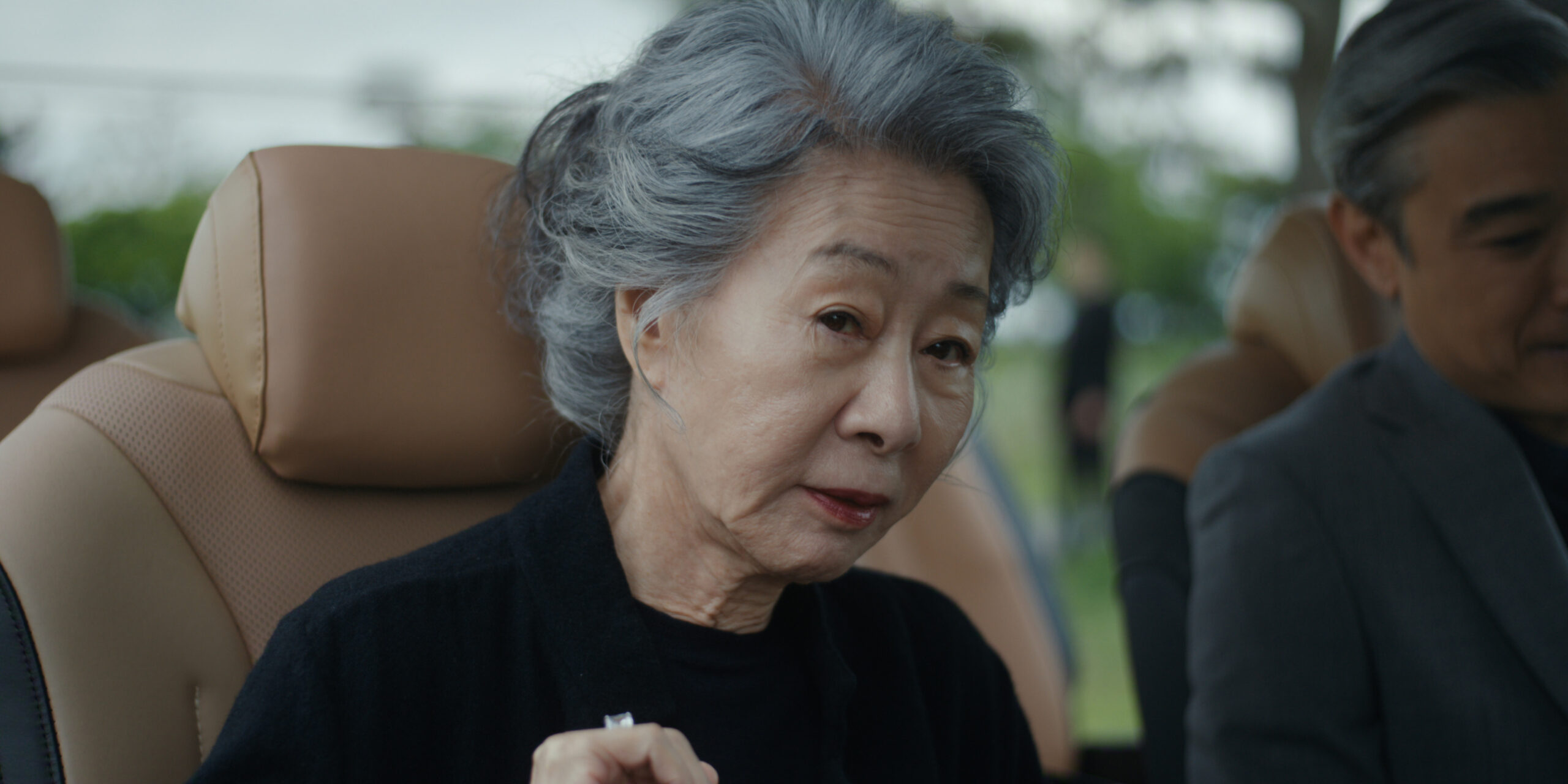 "Elderly woman with gray hair sitting in a car, wearing a black top, looking slightly toward the camera, with a man beside her. The car interior is tan, and greenery is visible outside the window."