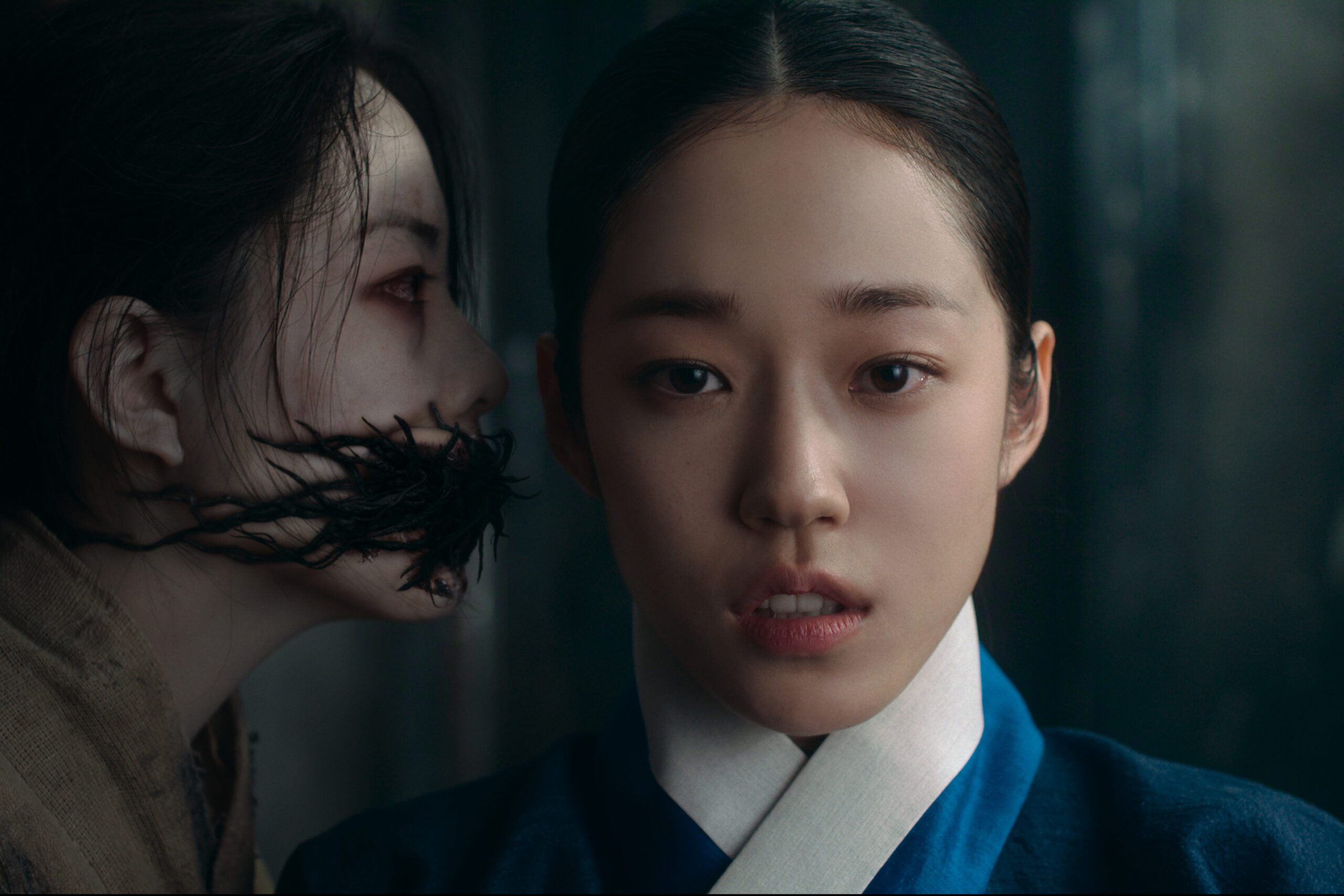 "Two women in traditional Korean attire, one with a fearful expression in the foreground and another with a disfigured, dark, vine-like mouth whispering behind her in a dimly lit, tense setting."